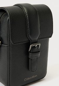 Fekete texturált bőrből készült Calvin Klein keresztpántos táska, elülső füllel és állítható csatos pánttal, fehér háttér előtt.