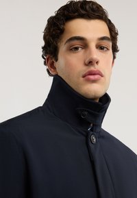 Marineblauer Mantel mit hohem Kragen, doppelter Knopfleiste und glatter Textur. Das Design zeichnet sich durch eine taillierte Silhouette und einen minimalistischen Stil aus.