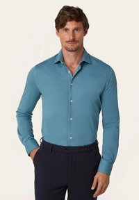 Langwerpige blouse met lange mouwen in teal stof met een gladde textuur. Heeft een puntkraag en zilveren knopen, gecombineerd met donkere broeken.