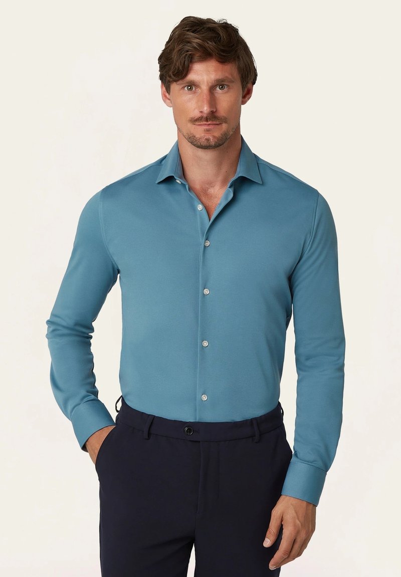 Langwerpige blouse met lange mouwen in teal stof met een gladde textuur. Heeft een puntkraag en zilveren knopen, gecombineerd met donkere broeken.