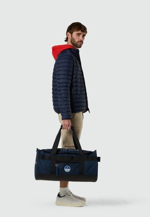 Homme portant une veste matelassée bleu marine à capuche rouge, short beige, baskets blanches, tenant un sac de sport bleu marine et noir avec un logo de boussole.