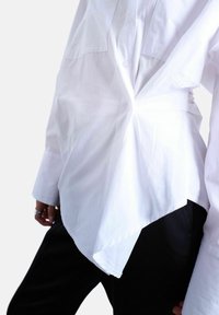 Personne portant une chemise blanche asymétrique boutonnée, rentrée dans un pantalon noir, montrant le design unique drapé de la chemise.