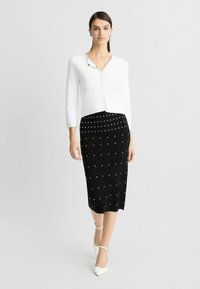 Blusa bianca aderente con maniche a tre quarti, abbinata a una gonna a matita nera con decorazioni a pois argentati e tacchi bianchi.