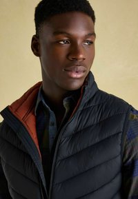 Junger schwarzer Mann, der eine schwarze gesteppte Weste über einem karierten Hemd trägt und leicht zur Seite blickt, vor einem neutralen Hintergrund.