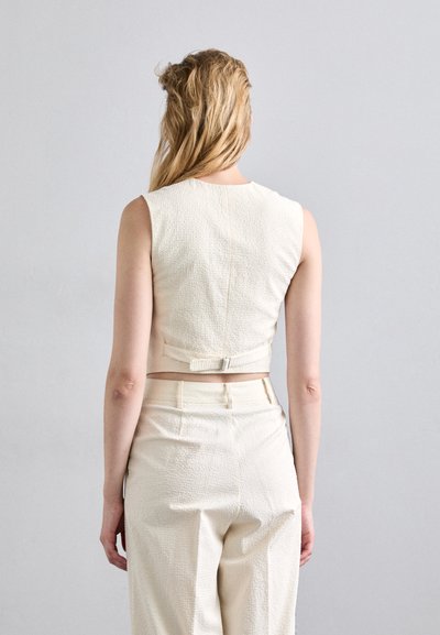 Top cropped en ivoire avec un tissu texturé, design sans manches et fermeture au dos. Associé à un pantalon taille haute assorti.
