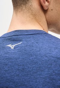 Κοντινό πλάνο στον αυχένα και τον ώμο ενός ατόμου που φοράει μπλε αθλητική μπλούζα με λευκό λογότυπο Mizuno στο πάνω μέρος της πλάτης.