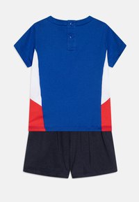 Body à manches courtes avec un haut bleu, des accents latéraux blancs et rouges, et un short marine foncé. Comprend des boutons-pression au dos pour la fermeture.