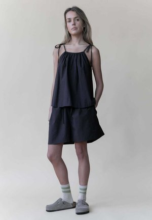 Zwarte gelaagde top met dunne bandjes en een losse pasvorm, gecombineerd met bijpassende shorts. Het model draagt grijze klompen en gestreepte enkelsokken.