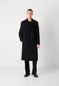 Un abrigo largo negro con un acabado texturizado, solapas anchas y bolsillos laterales, combinado con pantalones y zapatos negros, sobre un fondo neutral.