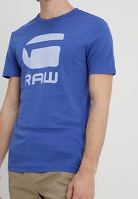 Mężczyzna w niebieskiej koszulce z krótkim rękawem z jasnoniebieskim abstrakcyjnym logo i napisem "RAW," w połączeniu z beżowymi spodniami na jednolitym tle.