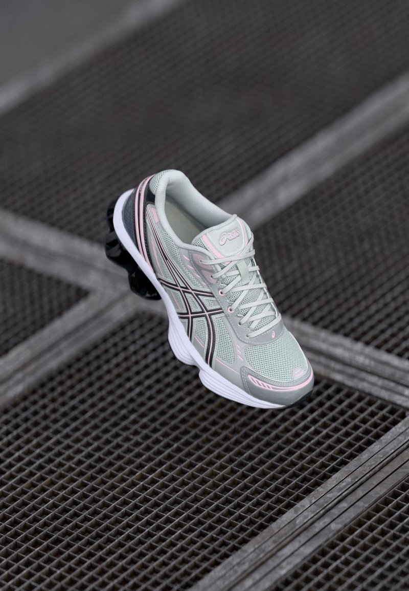 Chaussure de course gris clair et rose avec des accents noirs reposant sur une surface en grille métallique texturée, vue de dessus.