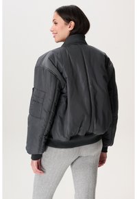 Eine Frau lächelt und trägt eine dunkelgraue gepolsterte Bomberjacke sowie eine hellgraue Rippstrickhose. Sie steht vor einem schlichten, hellen Hintergrund.