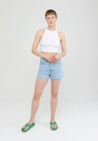 Weißes, kurzes ärmelloses Oberteil, hellblaue, hochtaillierte Shorts aus Denim mit fransigem Saum und grüne Flip-Flops, die vor einem neutralen Hintergrund stehen.