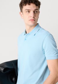 Gaiši zils polo krekls, kas izgatavots no kokvilnas, ar klasisku apkakli, īsām piedurknēm un diskrētu izciljuma logotipu. Nēsāts melns ādas soma.