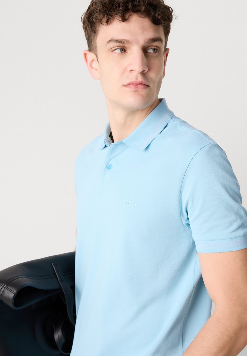 Lichtblauwe poloshirt van katoen, met een klassieke kraag, korte mouwen en een subtiele reliëflogo. Zwarte leren tas meegebracht.