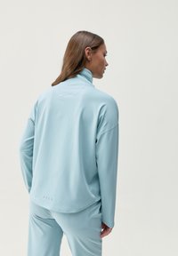Sudadera atlética de color azul claro con cuello alto, mangas largas y un corte relajado, con detalles de costura mínimos y una textura suave.