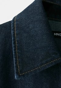 Colletto di camicia in denim blu scuro con cuciture marroni, che mostra la texture dettagliata del tessuto e parte di un'etichetta nera all'interno.