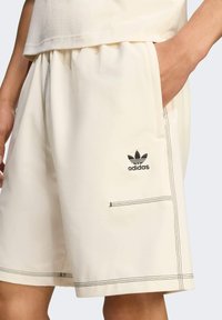 Kreminės spalvos šortai, pagaminti iš minkštos medžiagos su juodomis siūlėmis. Yra šoninės kišenės ir juodas Adidas logotipas šalia kairės kišenės.