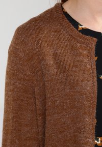 Brązowy dzianinowy kardigan z okrągłym dekoltem, wykonany z teksturowanej tkaniny z minimalnymi detalami przeszyć, noszony na czarnej kwiatowej sukience.