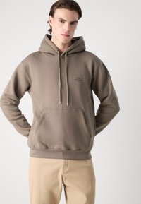 Sudadera marrón con bolsillo frontal, capucha con cordón y pequeño detalle de logo. La tela parece suave con una textura lisa. Combinada con pantalones beige.