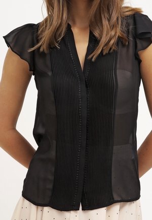 Blusa nera trasparente con maniche corte a volant, pieghe verticali e chiusura a bottoni frontale. Tessuto leggero e liscio.