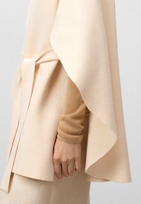 Beige gelaagd buitenkledingstuk met gedrapeerd ontwerp, vastgebonden in de taille. Daaronder een zachte, lichtbruine, aansluitende lange mouw top. Gladde textuur.