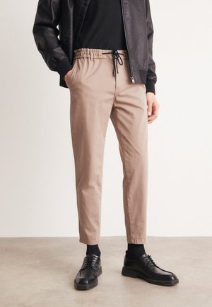 Pantaloni beige a sigaretta con vita elastica e dettaglio con cordoncino, abbinati a scarpe in pelle nere e calze nere, su sfondo neutro.