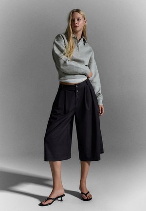 Szőke nő áll, szürke hosszú ujjú, rövidített pulóvert, fekete, széles szárú, gombos rövidített nadrágot és fekete magassarkú szandált visel.