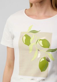 Wit t-shirt met korte mouwen, voorzien van een groene citroenafbeelding met bladeren, op een lichtbeige achtergrond en gouden tekstdetails aan de onderkant.