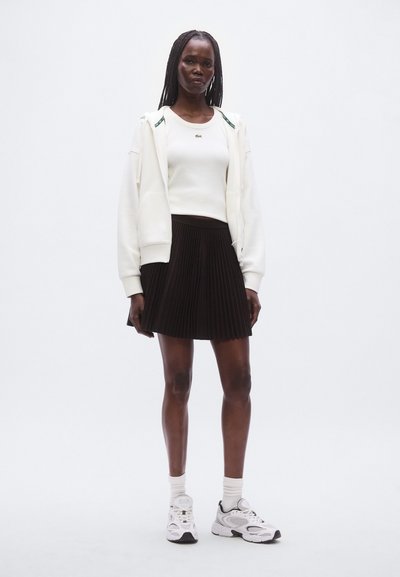 Sweat à capuche blanc zippé avec des accents verts, haut blanc près du corps, jupe plissée noire, baskets blanches et noires, debout sur un fond uni.