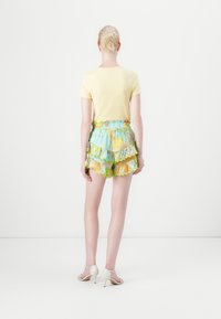 Keltainen t-paita yhdistettynä värikkäisiin kerroksellisiin shortseihin, joissa on sinisiä, vihreitä ja oransseja kuvioita; malli käyttää valkoisia korkokenkäsandaaleja.