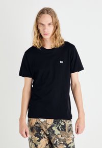 T-shirt nera in cotone con maniche corte, scollatura rotonda e un piccolo logo bianco "Lee" sul petto. Tessuto piatto, modello che indossa pantaloni mimetici.