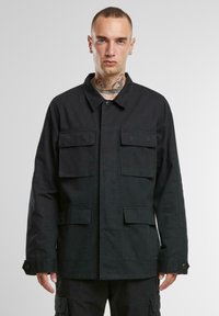 Svart arbetsjacka tillverkad av ett hållbart tyg, med krage, framknäppning, två bröstfickor och texturerade detaljer.