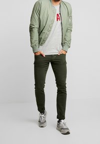 Mężczyzna w jasnozielonej kurtce bomberce, szarej koszulce z nadrukiem, ciemnozielonych dopasowanych spodniach i szarych tenisówkach stoi z rękami w kieszeniach.