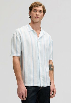 Mann trägt ein kurzärmeliges, weißes Hemd mit hellblauen vertikalen Streifen und dunkle Jeans, steht vor einem grauen Hintergrund.