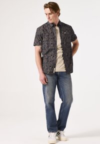 Korte mouwen, donker shirt met bladeren; beige T-shirt met ronde hals eronder, blauwe jeans en witte sneakers met groene accenten.