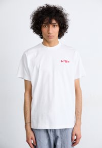 Camiseta blanca de algodón con mangas cortas, que presenta un pequeño texto gráfico rojo "Service Works" en la zona del pecho izquierdo.