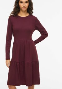 Robe à manches longues bordeaux en tissu côtelé. Un corsage ajusté se transforme en une jupe fluide à volants superposés. Encolure ronde, longueur mi-mollet.