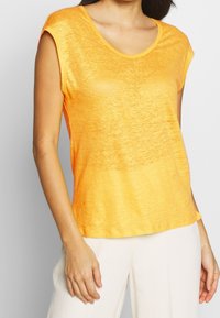 Oranžové tričko s krátkým rukávem a kulatým výstřihem, vyrobené z lehké látky, s jemně texturovaným povrchem. V kombinaci s světlými kalhotami.