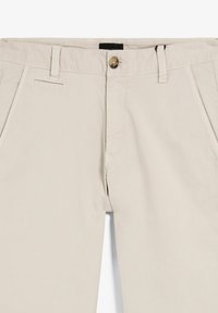 Chinos beige con frente plano, cierre de cremallera y un solo botón marrón. Presentan bolsillos laterales y un diseño limpio y ajustado.