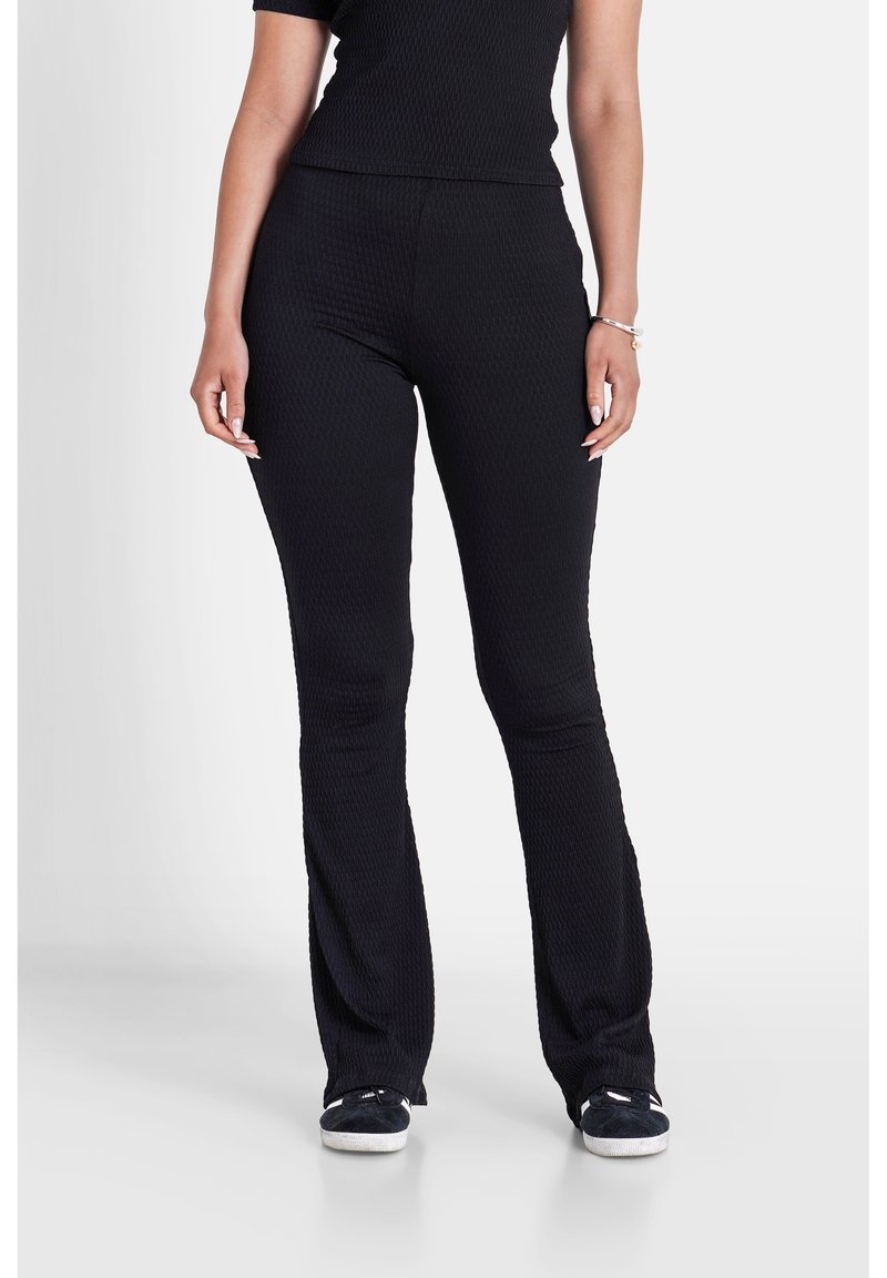 Femme portant un pantalon évasé noir texturé et un haut assorti, debout les mains détendues le long du corps et des baskets noires sur un sol blanc.