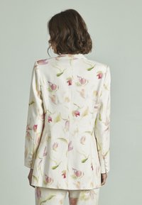 Blazer blanco con estampado floral en rosa y verde. Presenta un diseño estructurado, acentos de botones en las mangas y una parte trasera entallada.
