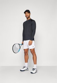 Ilga rankovė juoda palaidinė ir baltos šortai, laikant teniso raketę. Aksesuarai - juoda kepurė ir baltos sportinės bateliai su mėlynais akcentais.