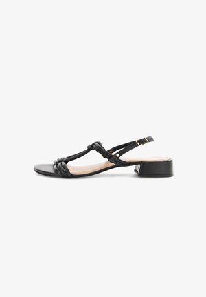 Sandalia negra de tacón bajo con tiras anudadas y correa con hebilla en el tobillo, punta abierta y tacón de forma cuadrada, mostrada sobre fondo blanco.