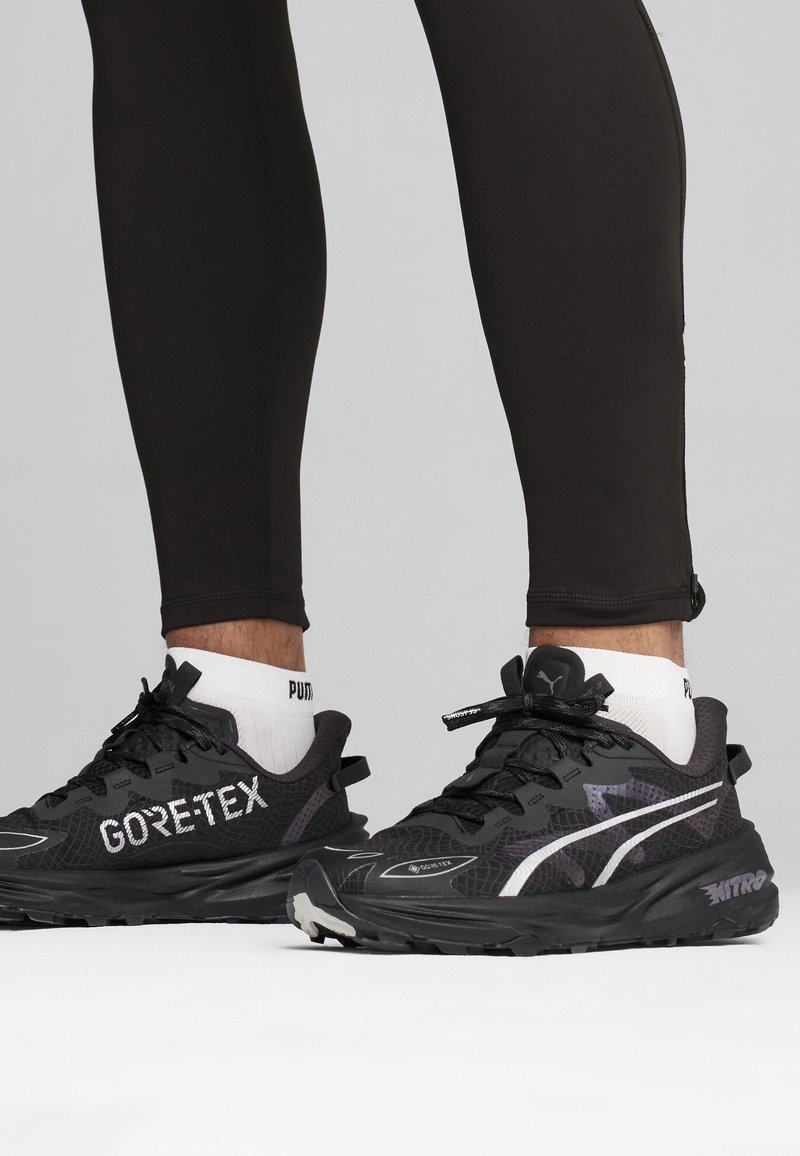 Zwarte GORE-TEX sneakers met een gestructureerde mesh bovenkant, witte accentstreep, robuuste zool en zwarte veters, gecombineerd met zwarte leggings en witte sokken.