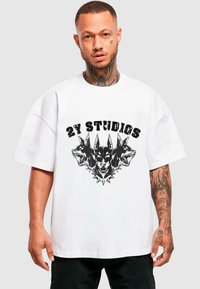 Bílá oversized tričko s grafikou tří černých psích hlav s hroty a textem "2Y STUIDOS" v tučném, opotřebovaném písmu.