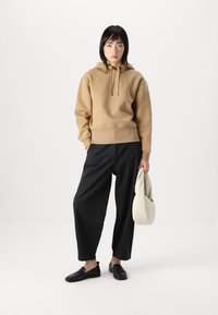 Beige mikina s kapucí a tkaničkami, volné černé kalhoty, černé nazouvací boty a světle béžová taška. Jednoduchý design s minimálními detaily.