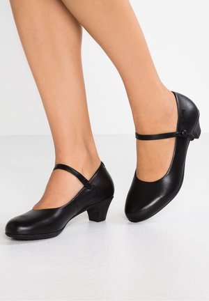 Kobieta nosząca czarne skórzane buty Mary Jane na niskim obcasie o zaokrąglonych czubkach, stojąca na białej podłodze.
