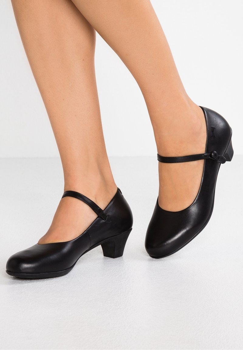 Femme portant des chaussures Mary Jane en cuir noir avec des talons bas et des bouts arrondis sur un sol blanc.