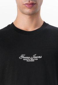 T-shirt en coton noir avec col rond, manches courtes, et logo "Guess Jeans" brodé en blanc avec "AMERICAN TRADITION CALIFORNIA" en dessous.